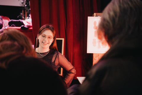 A smiling vendor wearing glasses and a sparkly top stands behind a table of Fluo stationery products at Oslo’s Supermarked. She’s talking to customers with red curtains in the background.