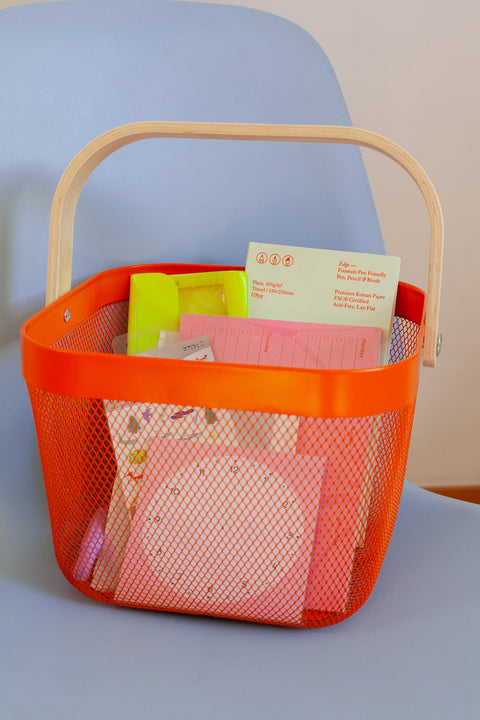 An orange shopping basket filled with colorful stationery like notebooks, planners, stickers and a neon yellow pencil case, placed on a light blue chair