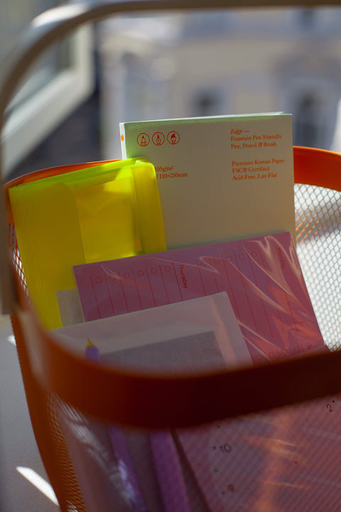 Close-up of an orange shopping basket in the sun, filled with colorful stationery like a weekly planner, a notebook, a pen and pencil, stickers and a neon yellow pencil case