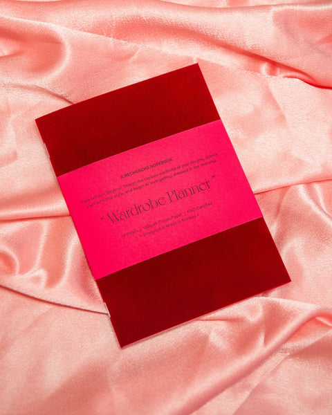 A capsule wardrobe planner made from velvet, deep red fabric, with a hot pink belly band around with product details. Placed on a rosy pink silk fabric.
