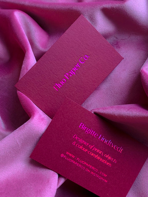 A deep wine-colored business card, debossed and printed with holographic pink foil. Lying on velvet pink fabric.