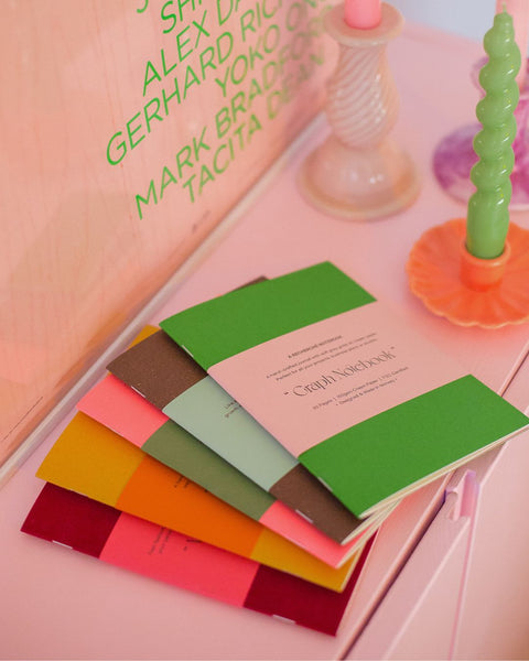 A set of five notebooks in assorted colors, lying on a pink surface with glossy, swirled candles to the right.
