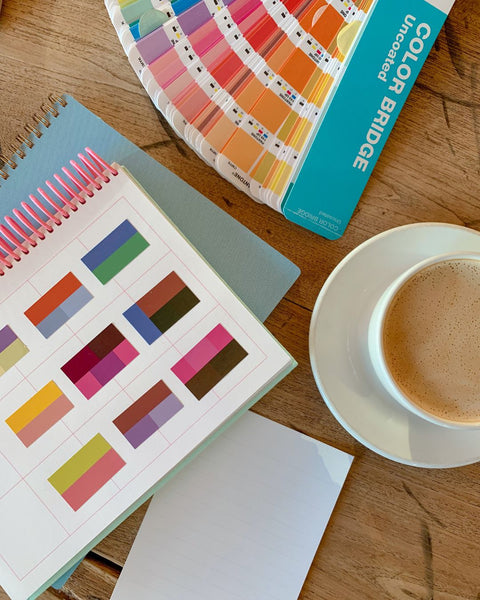A Pantone color bridge fanned on a wooden table, next to a notebook with printed color swatches stuck inside, and a cup of coffee.
