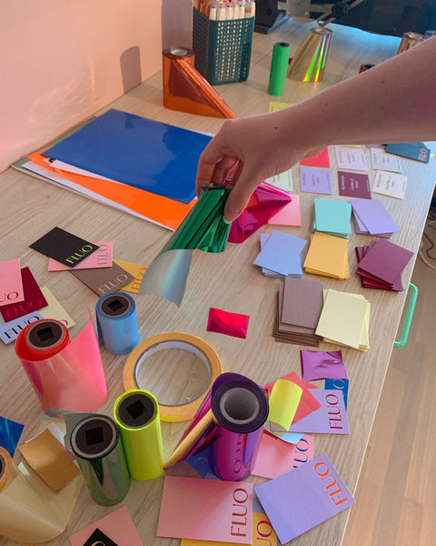 A person holding a roll of metallic green foil over a table that is scattered with other rolls of hot foil in assorted colors, and printed swatches with "Fluo" on them