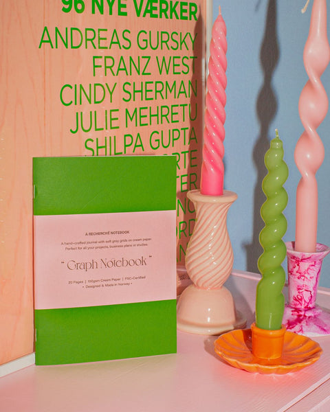 A green notebook standing in front of a contemporary pink and green typographic art print, with three glossy, colorful swirl candles to the right.