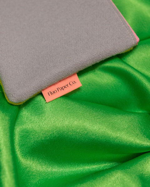 The corner of a grey wool pencil case, with a pink label that reads "Fluo Paper Co". Placed on a shiny green fabric.
