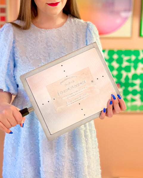 A person wearing a light blue dress, holding a metal printing plate to be used for hot foil printing on a KFS 3 platen foil press by Metallic Elephant.