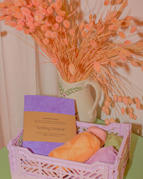 A knitting project journal placed in a color crate together with balls of yarn. Dried apricot-colored flowers in a cream vase in the background.