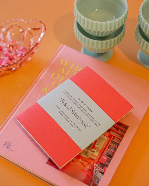 A coral notebook on top of a pink coffee-table book. Both placed on a pale orange table, with candy on the side.