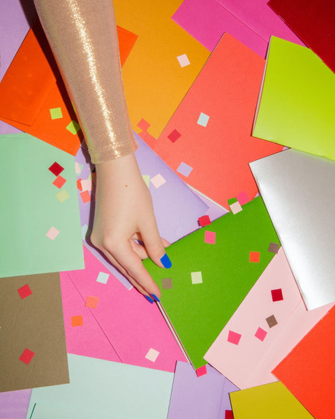 A bunch of colorful notebooks scattered on a table with confetti on top. An arm is reaching into the mix, grabbing one notebook. The person is wearing a shiny gold top and has bright blue fingernails