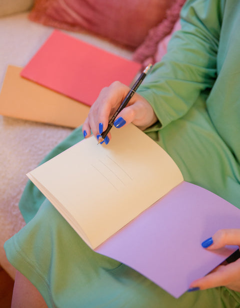 A person sitting on a sofa while wearing a green dress and blue nail polish, holding a grey mechanical pencil from Mark's Inc and a cream and purple-colored journal from Fluo