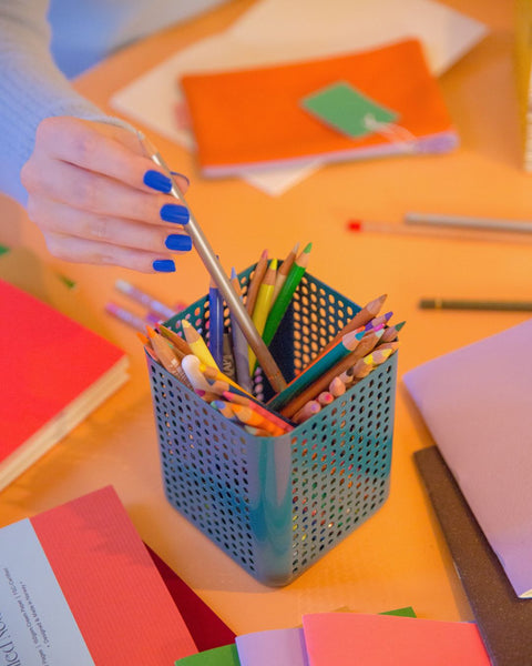 A hand with bright blue fingernails is reaching for a silver coloring pencil
