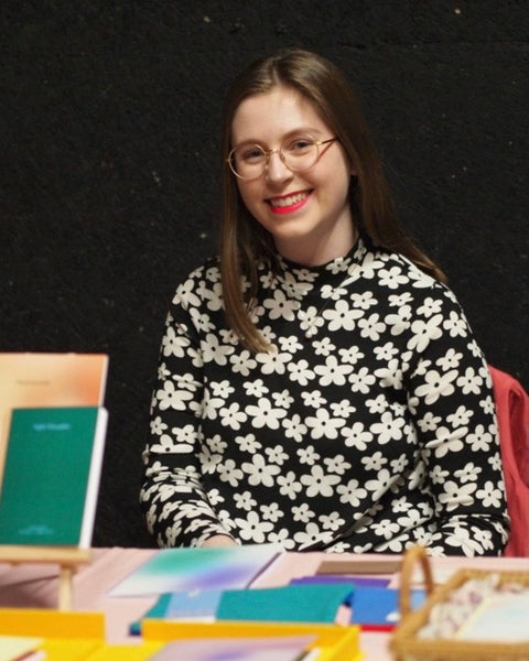 A person wearing a black dress with white flowers sitting behind a market stand table with notebooks and paper goods in the foreground