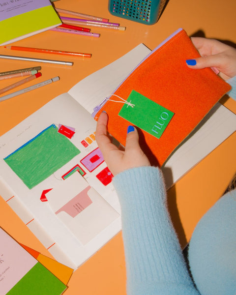 A person holding a pencil case. In the background: A sketchbook displaying product sketches of the same pencil case. Pencils laying around, everything placed on an apricot table.