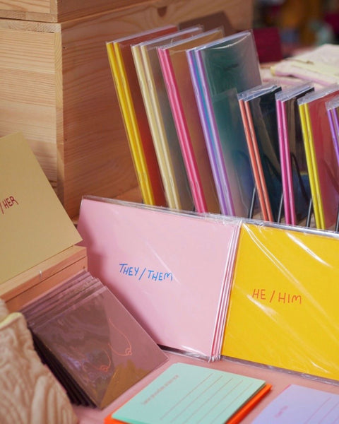 Art prints with foiled pronouns on them, displayed at a market stand table next to colorful notebooks and cards