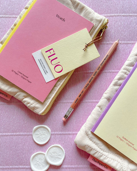 A flatlay of pencil cases and notebooks in pink, cream and yellow hues, with a pencil and some wax seals.