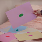 Hand holding a pink sheet of paper with a green oval in the center, depicting a blank mind map. Surrounded by similar sheets in different color combinations.