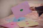 Hand holding a pink sheet of paper with a green oval in the center, depicting a blank mind map. Surrounded by similar sheets in different color combinations.