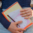 Person holding a notebook by Marjolein Delhaas. The front pocket of the notebook is clear and filled with Fluo memo sheets. The person is wearing a sparkly top, and also holds a mechanical pencil by Penco.
