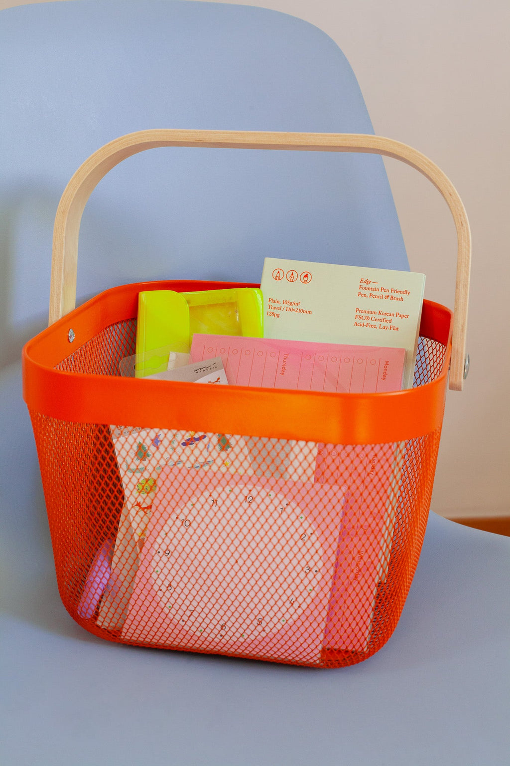 An orange shopping basket filled with colorful stationery like notebooks, planners, stickers and a neon yellow pencil case, placed on a light blue chair