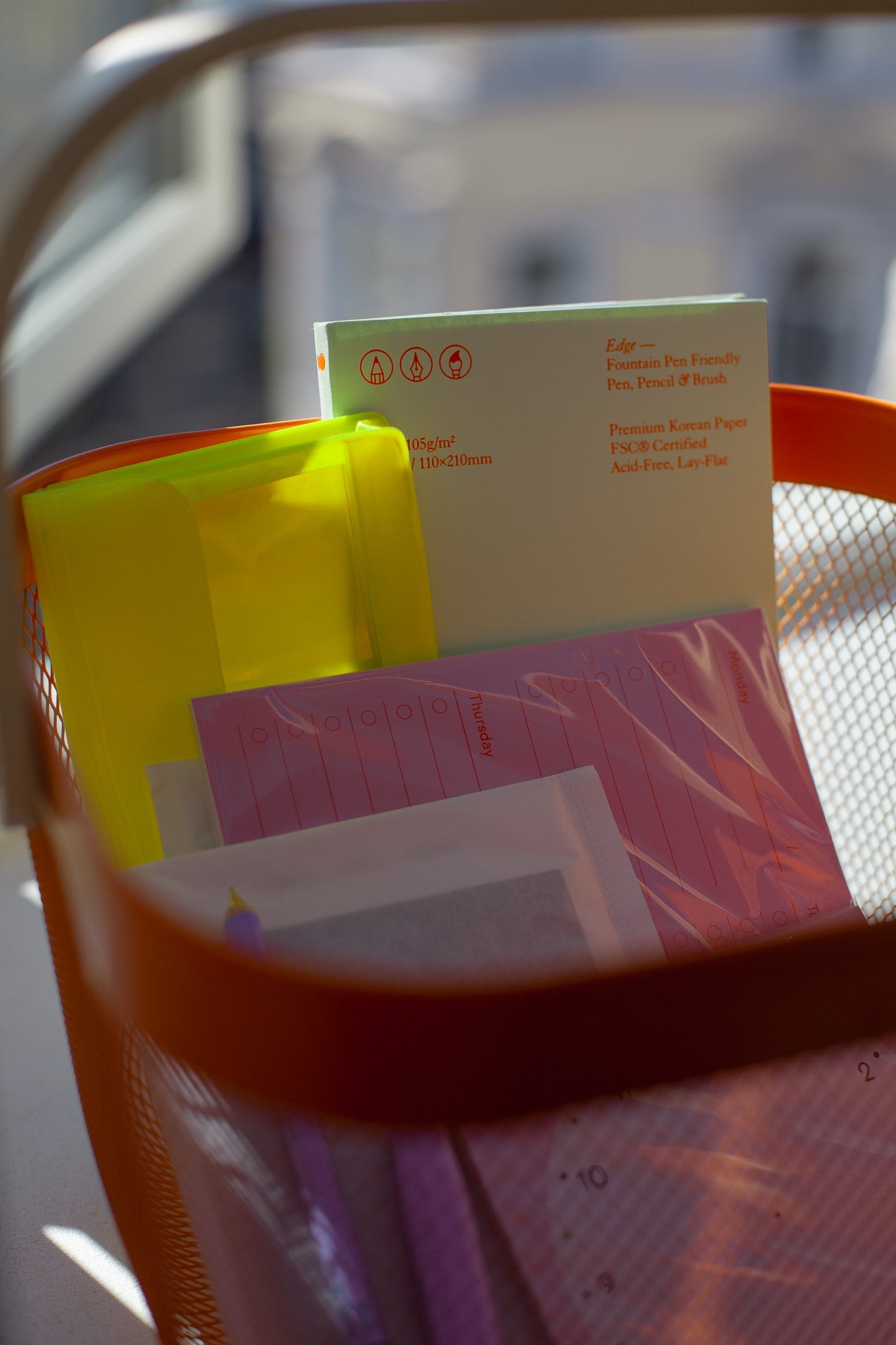 Close-up of an orange shopping basket in the sun, filled with colorful stationery like a weekly planner, a notebook, a pen and pencil, stickers and a neon yellow pencil case